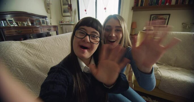 Cinematic shot of happy sister and teen girl with down making technology video call together to friends or family at home. Concept of conversation, handicapped, disability, media, love, friendship