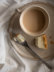 Morning cup of coffee and classic cheesecake on a beige ceramic plate on a soft white bed in the morning light