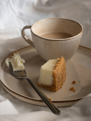 Morning cup of coffee and classic cheesecake on a beige ceramic plate on a soft white bed in the morning light