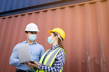 Team of logistic engineer man and woman wearing helmets with laptops holding hand and protective face mask standing outside containers area warehouse