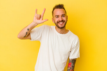 Young tattooed caucasian man isolated on yellow background showing a horns gesture as a revolution...