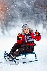 Boy in a red jacket and a hat with earflaps sledding down the hill in winter