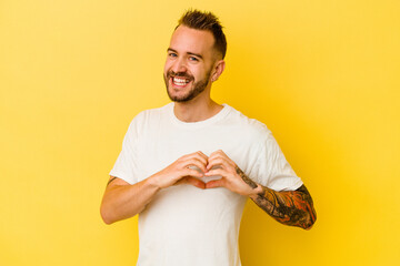 Young tattooed caucasian man isolated on yellow background smiling and showing a heart shape with...