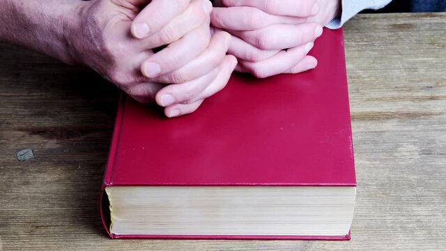 close-up of the hands of two men, young and old, folded in a prayer gesture on a thick family bible in a dark red cover, the concept of eternal values, the unity of people in faith
