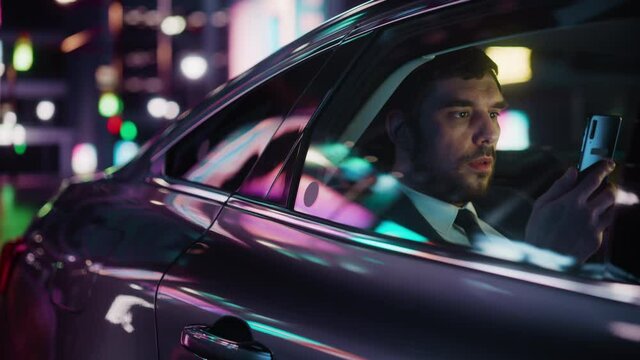 Handsome Businessman in a Suit Commuting from Office in a Backseat of His Car at Night. Entrepreneur Using Smartphone while in Transfer Taxi in Urban City Street with Working Neon Signs.