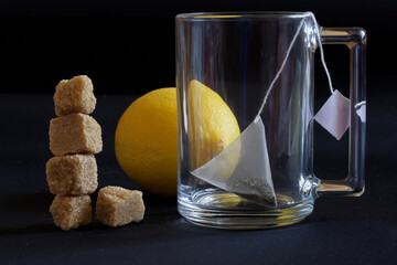 A glass transparent mug with a pyramid tea bag, fresh yellow lemon and a tower of brown cane unrefined sugar cubes. Dark background - obrazy, fototapety, plakaty