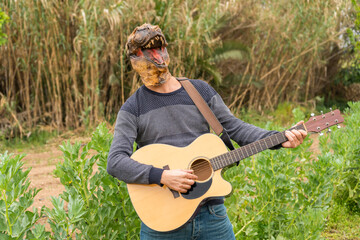 Happy man wearing dinosaur mask playing acoustic guitar outdoor in nature park at sunset.Humor...