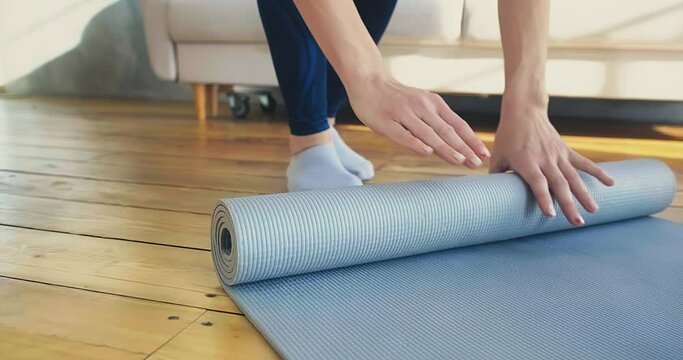 Young woman in socks rolls light grey rubber yoga mat by hands on wooden board floor in room after training closeup slow motion