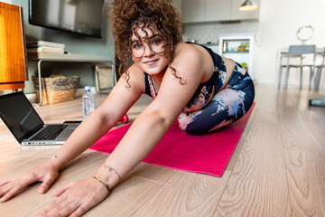 Young plus size woman at home doing exercise in front of open laptop, repeating instructions by...