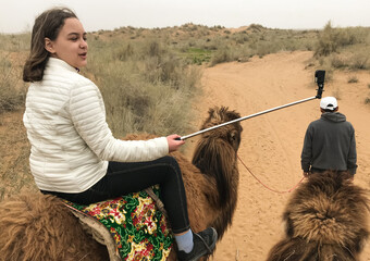 A teenage girl riding a camel in the desert with a selfie stick and a go pro camera as a travel...