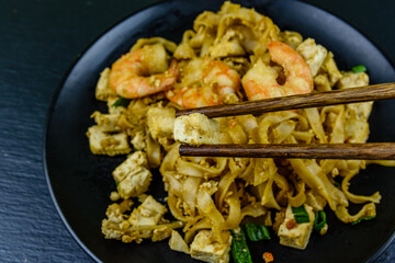 Black plate with pad thai on a slate board. Chopsticks with shrimp