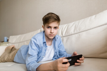 Child lies on the couch in his hands holding a phone, looks into the camera.