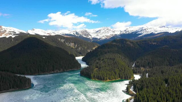 Aerial view of a frozen mountain lake surrounded by forest and snowy mountains in spring season. Lake covered by a thick layer of ice. Spring in the mountains. Turquoise crystal water.