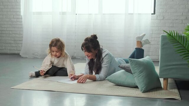 Mom and cute little daughter lying on carpet on the floor at home drawing on paper together, spending time painting with colorful pencils.
