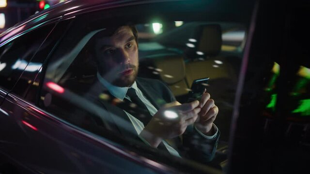 Handsome Businessman in a Suit Commuting from Office in a Backseat of His Luxury Car at Night. Entrepreneur Using Smartphone while in Transfer Taxi in Urban City Street with Working Neon Signs.