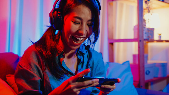 Happy asia girl gamer wear headphone competition video game online with smartphone excited talk with friend sit on couch in colorful neon lights living room at home, Home quarantine activity concept.