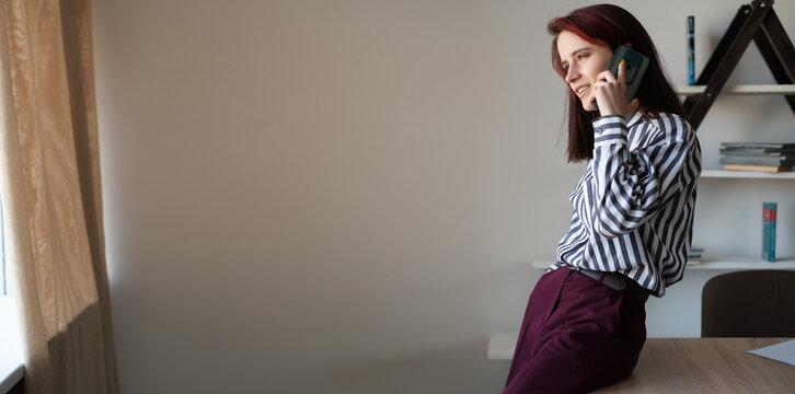 A Young Caucasian Girl, 20 Years Old, A Teacher Or A Consultant, Stands At A Table In The Office And Talks On The Phone. Remote Work Or Education Concept With Gadgets.