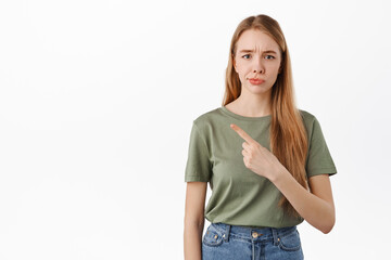 Obraz premium Not so sure. Skeptical young woman smirk and frown from doubts, pointing left at something average, judging bad promo deal, standing over white background