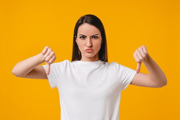 Sad, angry mistrusted young hispanic woman showing thumbs down dislike isolated over yellow...