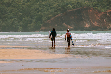 Para surferów, dziewczyna i chłopak idący z deskami surfingowymi po plaży na tle oceanu i klifu.