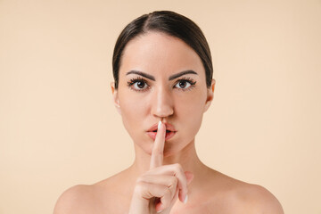 Fototapeta premium Pretty caucasian young woman making silent gesture with bare shoulders naked with clean skin isolated over beige background. Top secret for cosmetology, beautification, spa concept