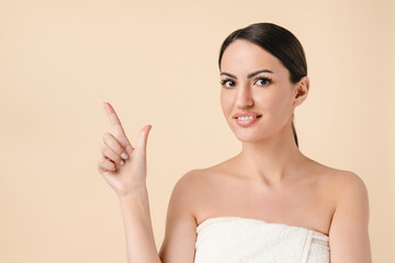 Caucasian beautiful young woman in bath spa towel pointing at copy space isolated over beige background. Pretty young white woman ready for beautification and spa, cosmetology, make up concept.