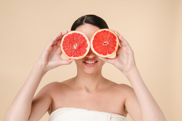 Young attractive caucasian woman holding grapefruits near her face isolated over beige background. White woman in spa towel waiting for spa and body care procedures. Pampering, moisturizing effect