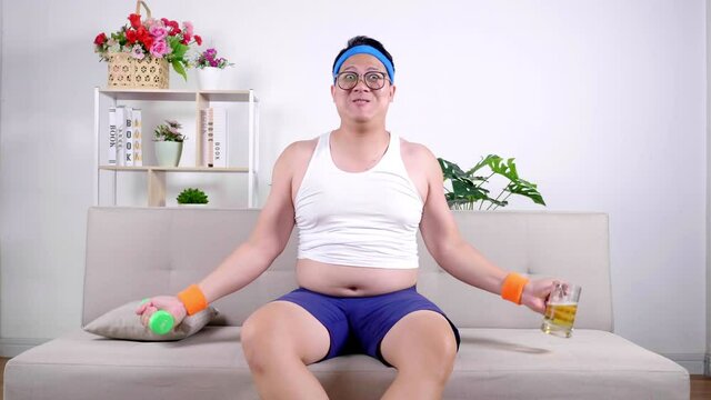 Funny fat Asian guy in sportswear lifting dumbbell weight training with enjoy drinking beer for refresh energy. Happy overweight man workout and sitting on couch at living room while quarantine