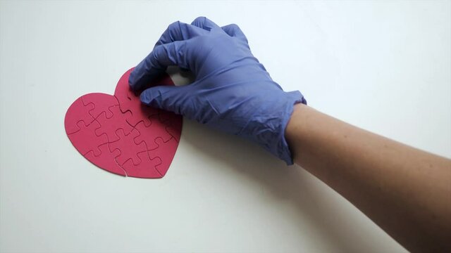 Woman's hands in blue medical latex gloves places one piece of puzzle on the missing part to red heart shaped jigsaw on white table. Top view