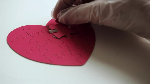 Extreme close up. Woman's hands in white medical latex gloves places one piece of puzzle on the missing part to red heart shaped jigsaw on white table. Top view