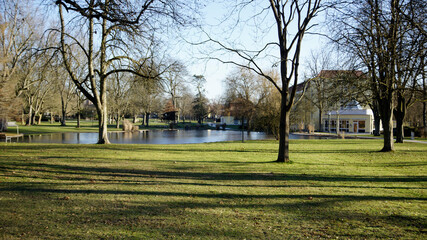 Obraz premium Jugendstil Kurhaus vor Seenfläche und kahlen Bäumen im Kurpark in Bad Hersfeld im Frühling mit Sonne ohne Menschen