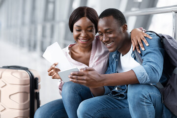 Waiting For Flight. Happy Black Couple Using Smartphone In Airport Terminal