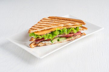 Single sandwich consisting of cucumber, slice of meat and cheese, lettuce and tomatoes between slices of grilled bread served on plate on white wooden background at kitchen. Horizontal image