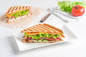 Homemade healthy sandwich made of cucumber, slice of meat and cheese, lettuce and tomatoes between slices of grilled bread served on plate on white wooden background with ingredients. Horizontal