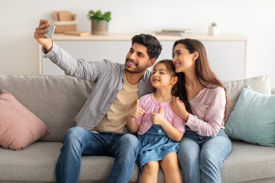 Family selfie. Happy arab man taking photo with his wife and little daughter, sitting on sofa at home