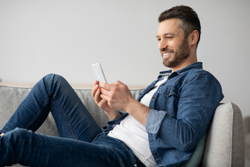 Happy man resting on couch at home, using modern smartphone