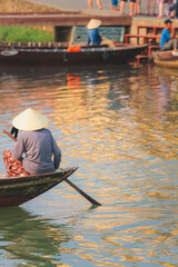 Obraz premium A local Vietnamese woman wearing a traditional conical leaf hat (non la) paddles in the Thu Bon River in the quaint old town village Hoi An, Vietnam.