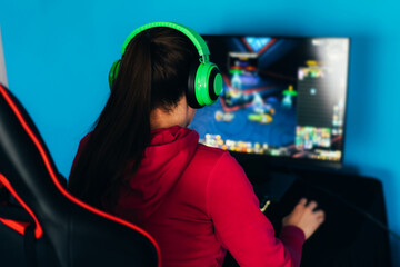 The gamer sits on a gaming chair and plays computer games. The player has green headphones on his head