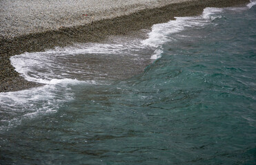The wave runs onto the pebble shore and rolls back with white foam. beautiful natural background. A foamy sea wave runs along the rocky shore. Sea foam, pebbles