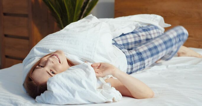 Happy morning. Full length view of the handsome ginger girl peeking up under the blanket at her bed and looking at the camera with pleasure face. Happy woman enjoying of her morning at home