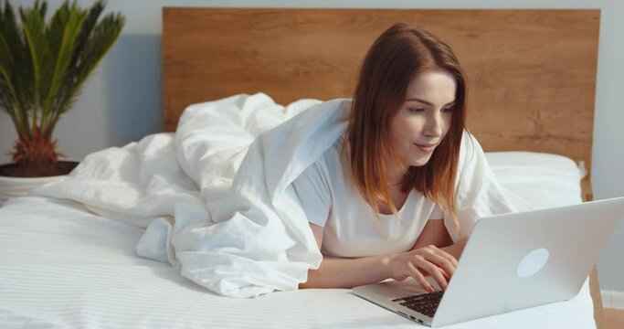 Full length view of the focused woman using laptop at home, looking at screen, chatting, reading or writing email. Girl laying at the bed, while doing homework or working on research project online