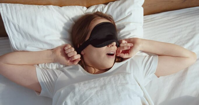 Ginger cute woman laying at the bed and taking off her face sleep mask while waking up at the morning at her cozy bedroom. Woman laying at the white sheets with great mood