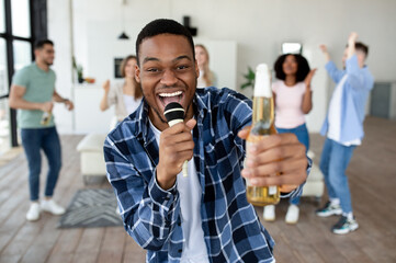 Cheerful black guy with bottle of beer and microphone singing karaoke with friends at home