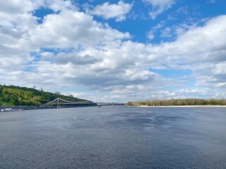 Ukraine Kiev city landscape. Dnieper river calm blue water at summer sunny day. Summertime, tranquility, relax. Scenic natural landscape. Spectacular cloudy sky. Pedestrian bridge over the river. 