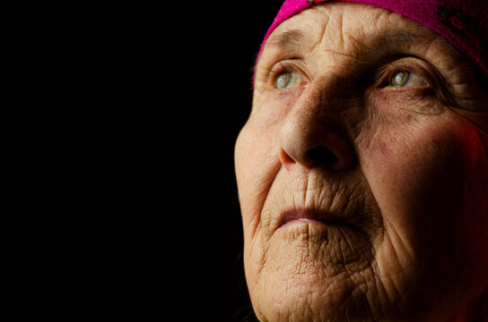 The face of an old grandmother close up. Close-up of the wrinkled face of an old grandmother. Portrait photo of an elderly woman in the dark. Seniors concept. Old grandmother. Old woman looking up