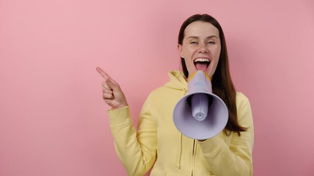 Excited cute young brunette female screaming in megaphone pointing index finger aside up, wears yellow hoodie, posing isolated on pink background with copy space. People emotion lifestyle concept