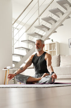 Calm Healthy Serene Old Middle Aged Senior Tattooed Man Meditating With Eyes Closed Relaxing Sitting In Yoga Pose In Living Room At Home Doing Breathing Exercises, Feeling No Stress, Peace Of Mind.