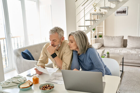Worried old senior couple checking bank documents at home. Serious mature retired family reading paper bills, calculating taxes, planning retirement finances, household expenses, having money problems