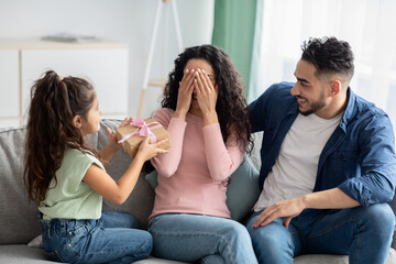 Portrait of cute little girl surprising her mom, giving present at home