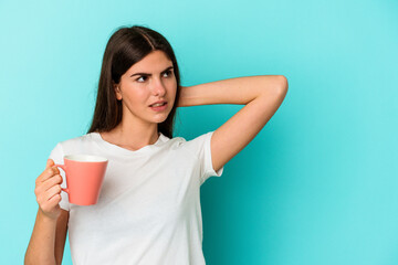 Young caucasian woman holding a mug isolated on blue background touching back of head, thinking and making a choice.